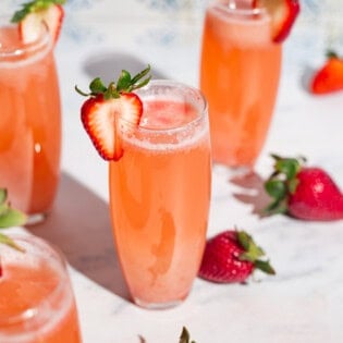 Four strawberry bellinis in glasses garnished with strawberry slices surrounded by whole strawberries.