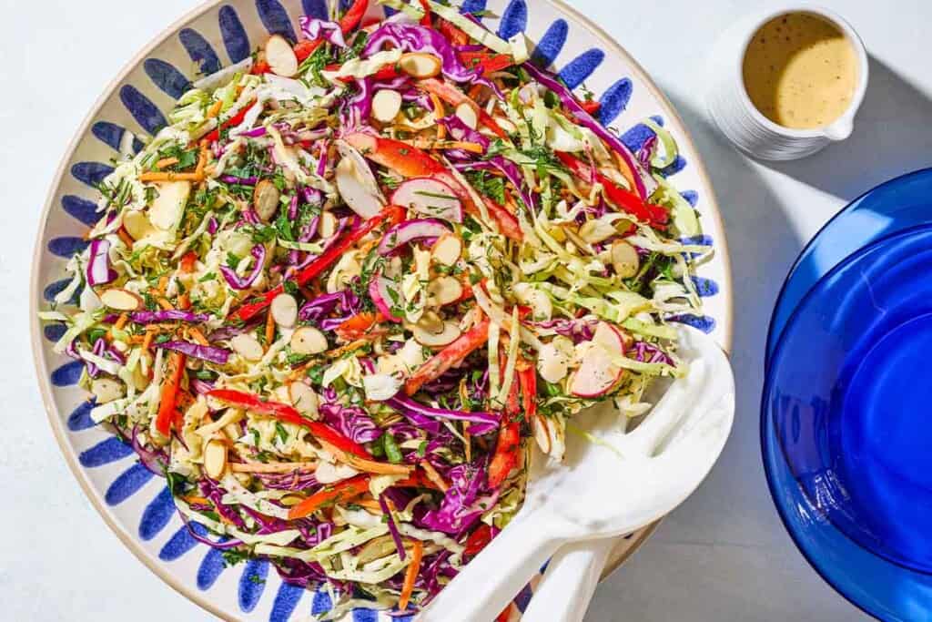 An overhead photo of cabbage salad in a serving bowl with serving utensils. Next to this is a small container of the dressing and a stack of 2 plates.