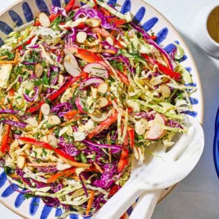 An overhead photo of cabbage salad in a serving bowl with serving utensils. Next to this is a small container of the dressing and a stack of 2 plates.