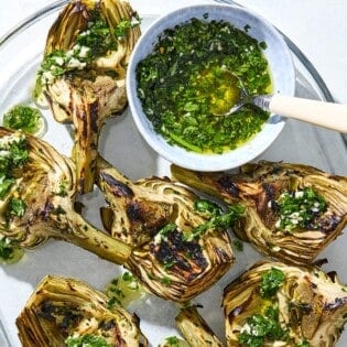 Grilled artichoke quarters drizzled with marinade on a serving platter along with some of the marinade in a bowl with a spoon.