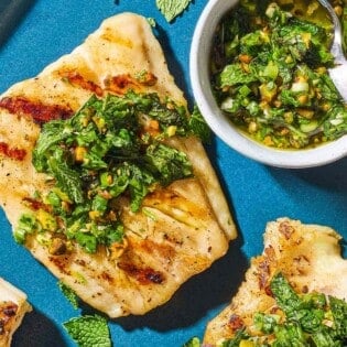 An overhead photo of 2 grilled cod filets topped with pistachio-herb salsa on a serving platter along with a small bowl of the salsa and a spoon.