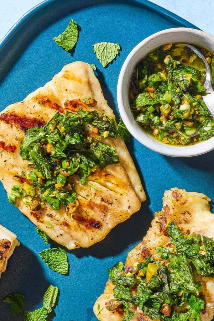 An overhead photo of 2 grilled cod filets topped with pistachio-herb salsa on a serving platter along with a small bowl of the salsa and a spoon.