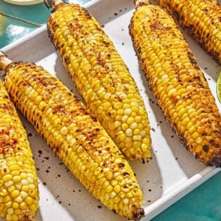 An overhead photo of 5 grilled corncobs on a serving platter with lime half. Next to this is a small bowl of Aleppo pepper.