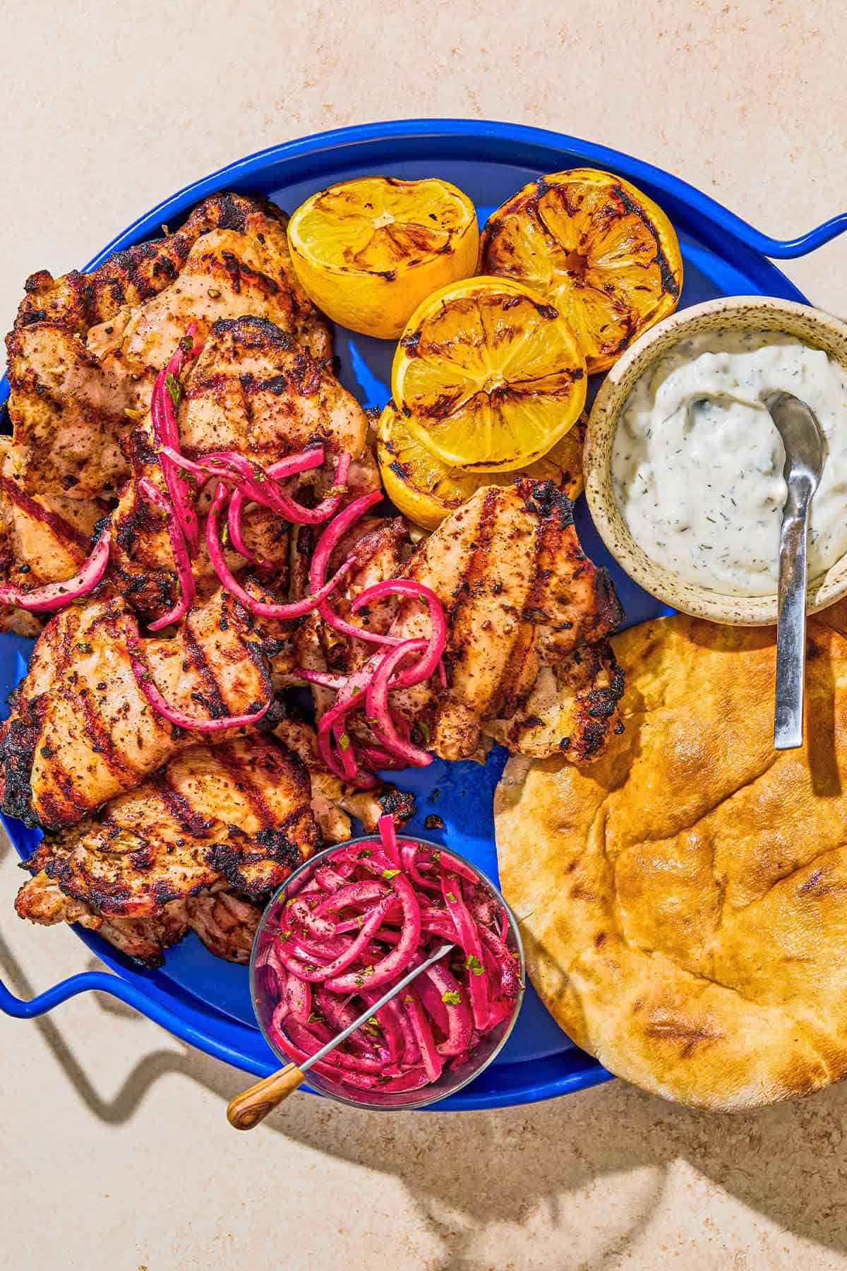 An overhead photo of grilled sumac chicken thighs on a serving platter with pickled red onions, grilled lemon halves, tzatziki sauce, and pita bread.