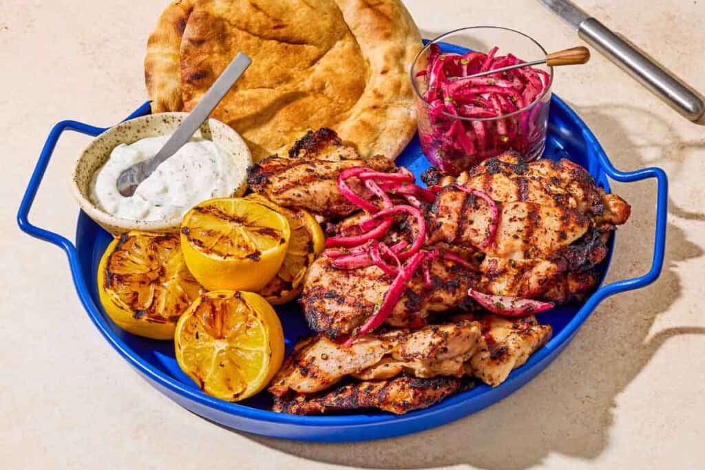 Grilled sumac chicken thighs on a serving platter with pickled red onions, grilled lemon halves, tzatziki sauce, and pita bread.