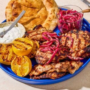 Grilled sumac chicken thighs on a serving platter with pickled red onions, grilled lemon halves, tzatziki sauce, and pita bread.