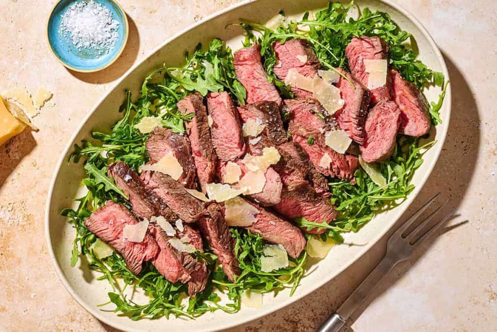 An overhead photo of beef tagliata topped with shaved parmesan on a bed of arugula on a serving platter. Next to this is a small bowl of salt and a serving fork.