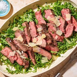 An overhead photo of beef tagliata topped with shaved parmesan on a bed of arugula on a serving platter. Next to this is a small bowl of salt and a serving fork.
