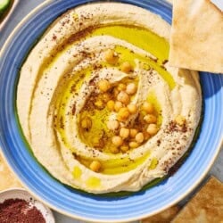 An overhead photo of hummus in a bowl topped with olive oil, chickpeas and sumac. Next to this is a small bowl of sumac, pieces of pita bread, and a bowl of sliced cucumber.