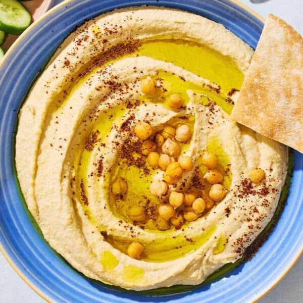 An overhead photo of hummus in a bowl topped with olive oil, chickpeas and sumac. Surrounding this is a bowl of sliced cucumber and pieces of pita bread.