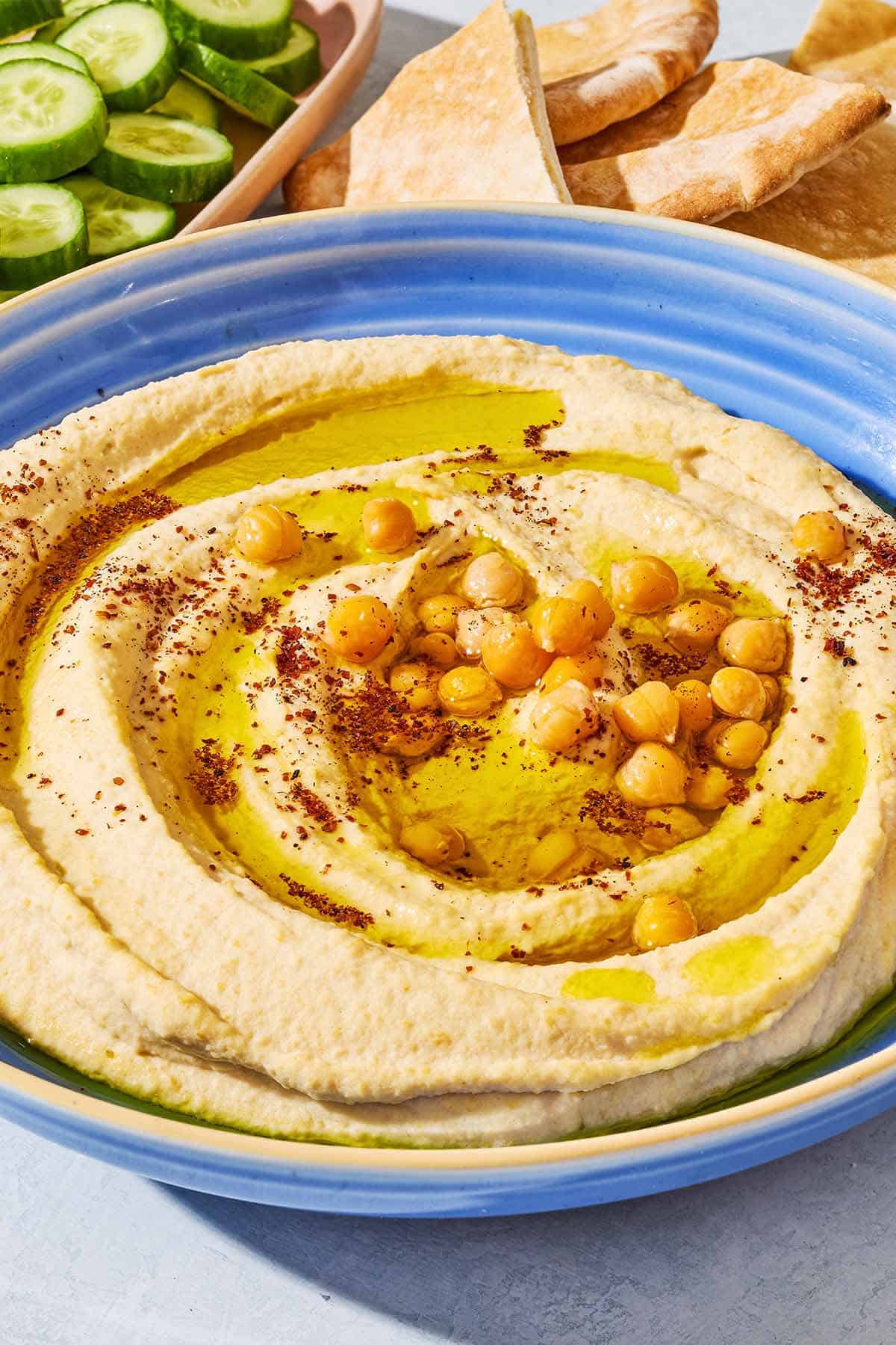 A close up of hummus in a bowl topped with olive oil, chickpeas and sumac. In the background is a bowl of sliced cucumber and pieces of pita bread.