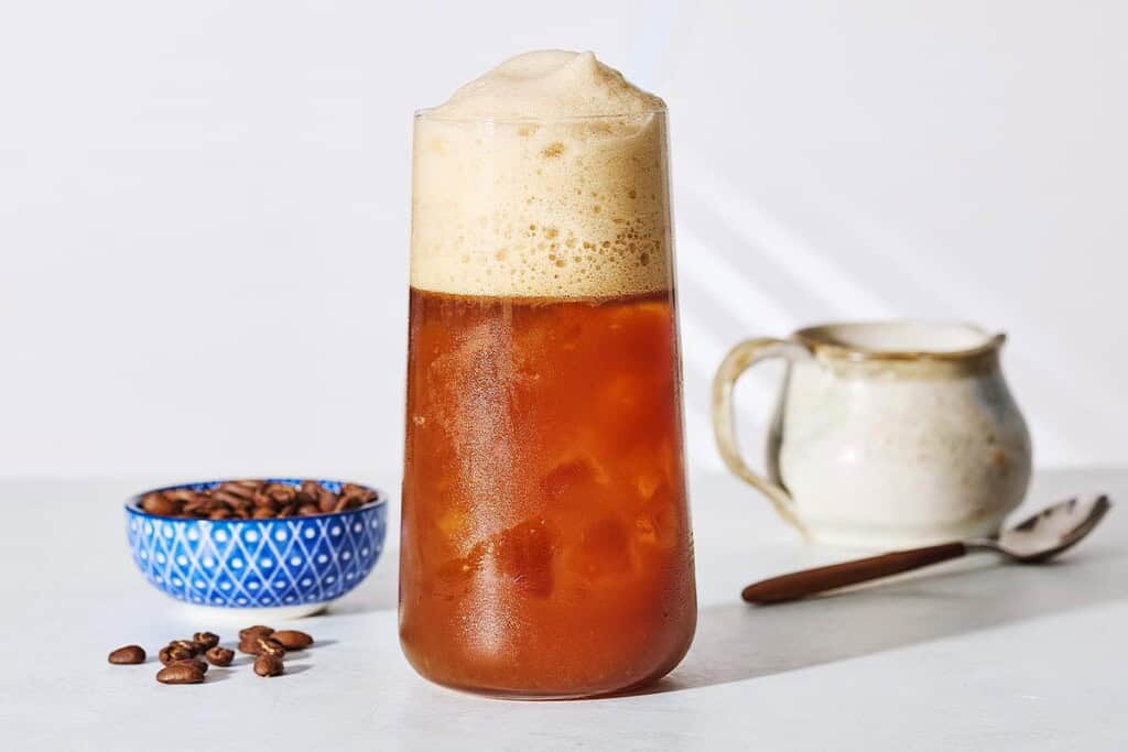 A close up of Freddo Espresso in a glass next to a bowl of coffee beans, a small pitcher and a spoon.