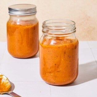 Two jars of roasted tomato sauce, one of them with a lid on it. There is a serving spoon next to the jars.