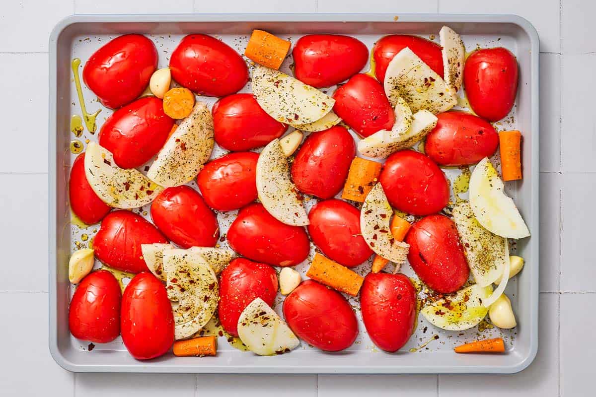 An overhead photo of seasoned, uncooked onions, tomatoes, carrots and garlic on a baking sheet.