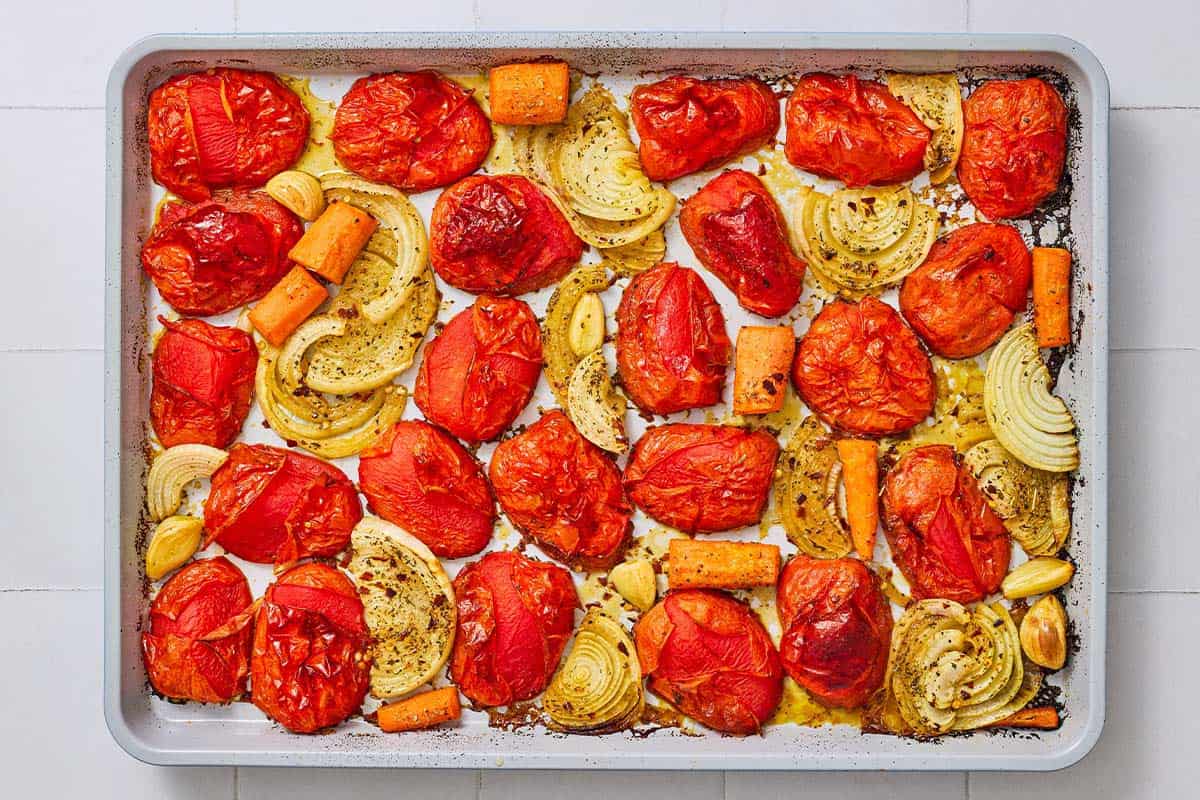 An overhead photo of roasted onions, tomatoes, carrots and garlic on a baking sheet.