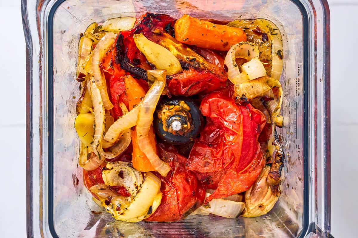 An overhead photo of roasted onions, tomatoes, carrots and garlic in a blender just before being mixed together.