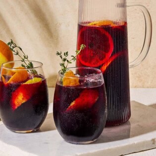 A close up of a pitcher non alcoholic sangria next two glasses of the sangria garnished with sprigs of fresh thyme and orange and lemon peels on a marble serving tray. Behind this is a bowl of oranges.