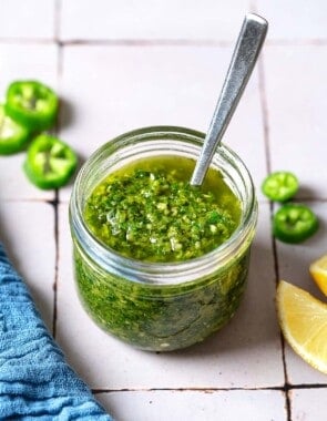 A close up of zhoug in a jar with a spoon. Next to this is a kitchen towel, slices of jalapeno and 2 lemon wedges.