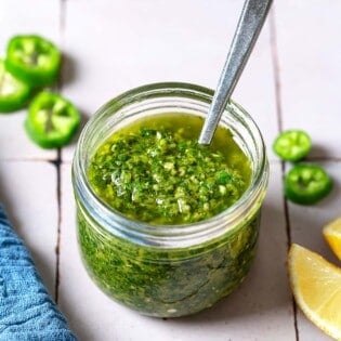 A close up of zhoug in a jar with a spoon. Next to this is a kitchen towel, slices of jalapeno and 2 lemon wedges.