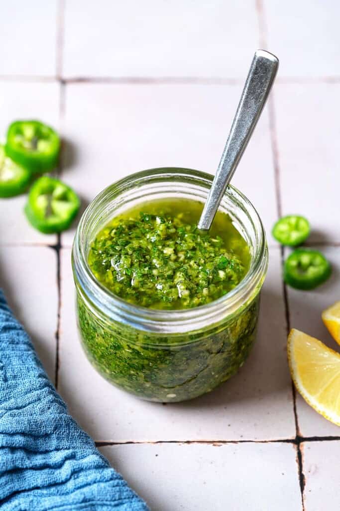 A close up of zhoug in a jar with a spoon. Next to this is a kitchen towel, slices of jalapeno and 2 lemon wedges.
