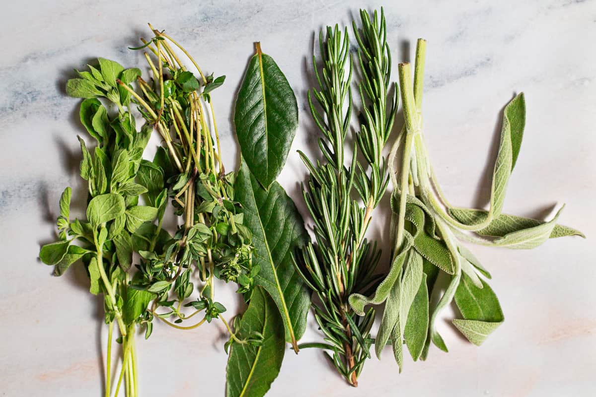 Woody herbs in groups on a white marble background, showing oregano, thyme, bay leaves, sage, and rosemary with little space in between each.