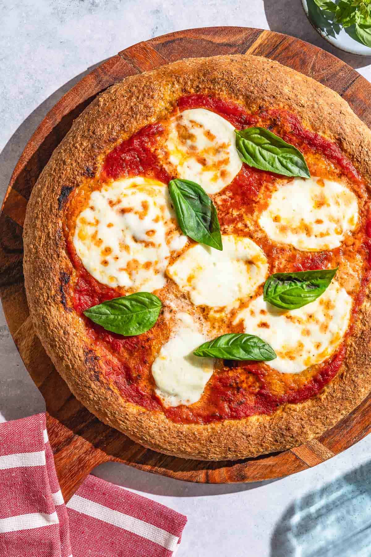 An overhead photo of a pizza with a whole wheat pizza crust on a wooden serving platter with a kitchen towel.