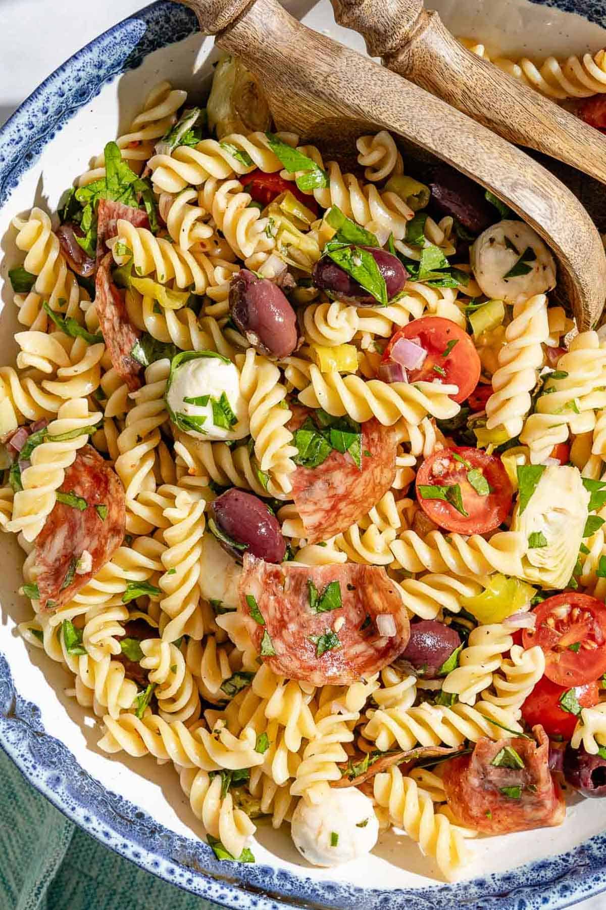 Close up of Italian pasta salad in a bowl with two wooden serving spoons. You can see the variety of textures and color from the cooked pasta, mozzarella, tomatoes, fresh herbs, olives, salami, and artichoke hearts.