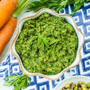 Carrot top pesto in a bowl next to a bunch of carrots and bowls of salt and pistachios.