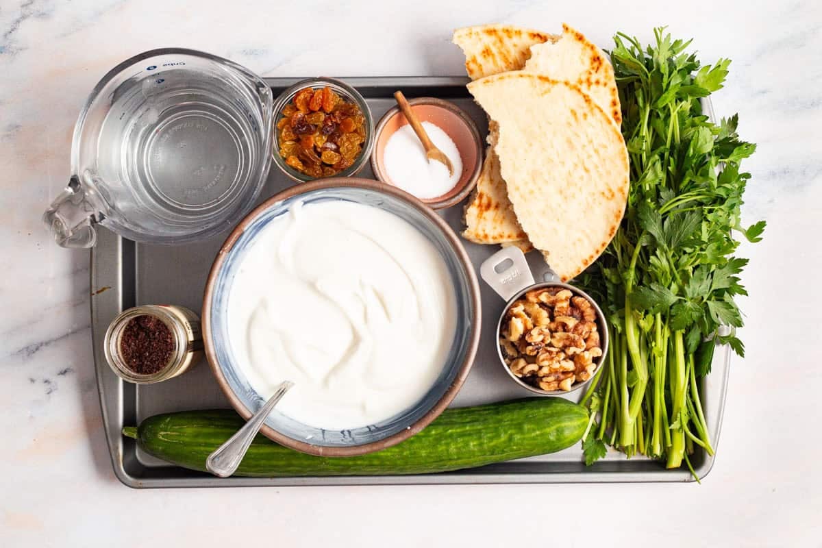 Ingredients for cucumber soup including cucumber, yogurt, walnuts, water, parsley, golden raisins, sumac, salt and some pita bread.