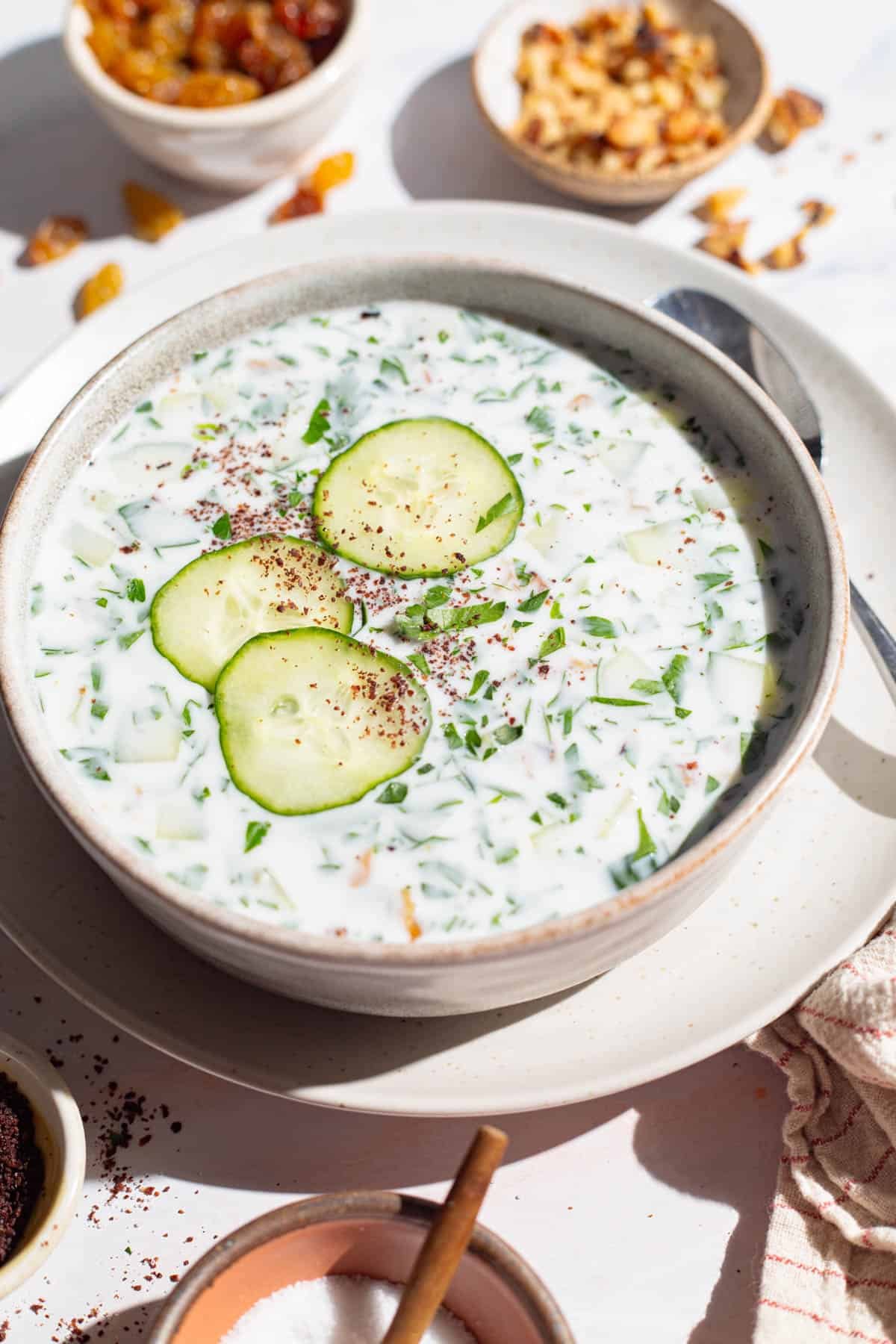 A bowl of cucumber soup garnished with sumac, parsley and cucumber slices on a plate with a spoon. Next to this is a kitchen towel and bowls of salt, golden raisins and walnuts.
