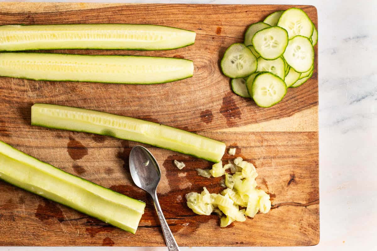 An overhead photo of cucumber slices and 4 cucumber quarters on a cutting board with a spoon. The seeds have been scooped out of one of the cucumber pieces with the spoon.