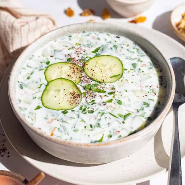 A bowl of cucumber soup garnished with sumac, parsley and cucumber slices on a plate with a spoon. Next to this is a kitchen towel and bowls of sumac and walnuts.