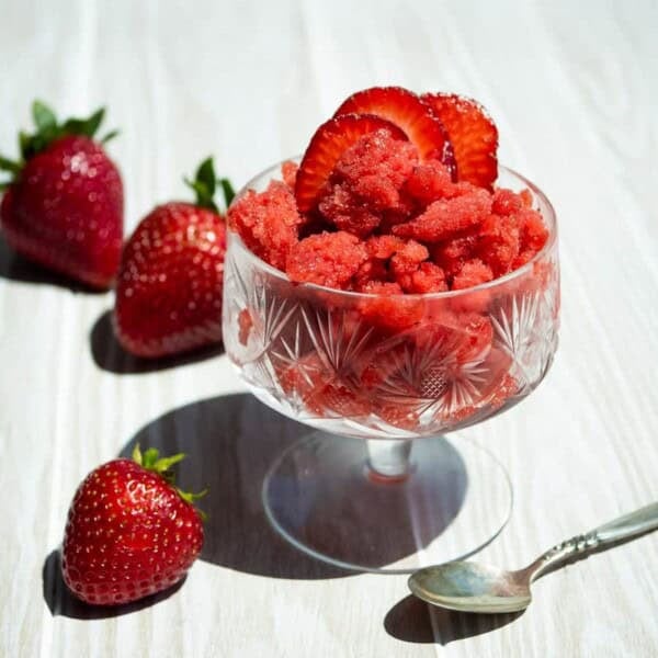 A strawberry granita in a glass garnished with strawberry slices. Next to this is a spoon and 3 strawberries.