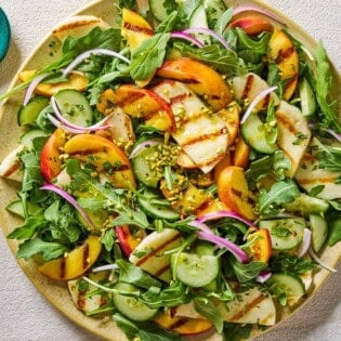 Halloumi and peach salad on a platter next to small bowls of chopped mint leaves and crushed pistachios.
