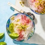 An overhead photo of the faloodeh in a bowl topped with the cherry syrup, chopped pistachios and lime wedges. Next to this is a spoon, 2 lime wedges, a bowl of the pistachios and another bowl of the faloodeh.