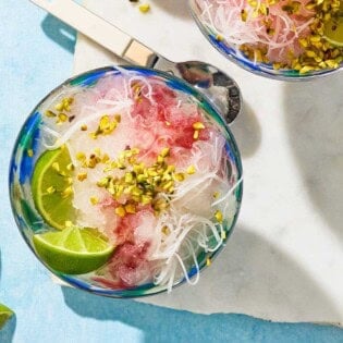 An overhead photo of the faloodeh in a bowl topped with the cherry syrup, chopped pistachios and lime wedges. Next to this is a spoon, 2 lime wedges, and another bowl of the faloodeh.