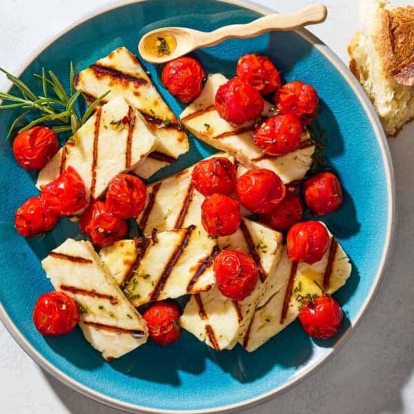 An overhead photo of grilled halloumi with blistered cherry tomatoes, sprigs of rosemary and a small spoon on a serving platter next to some chunks of crusty bread.