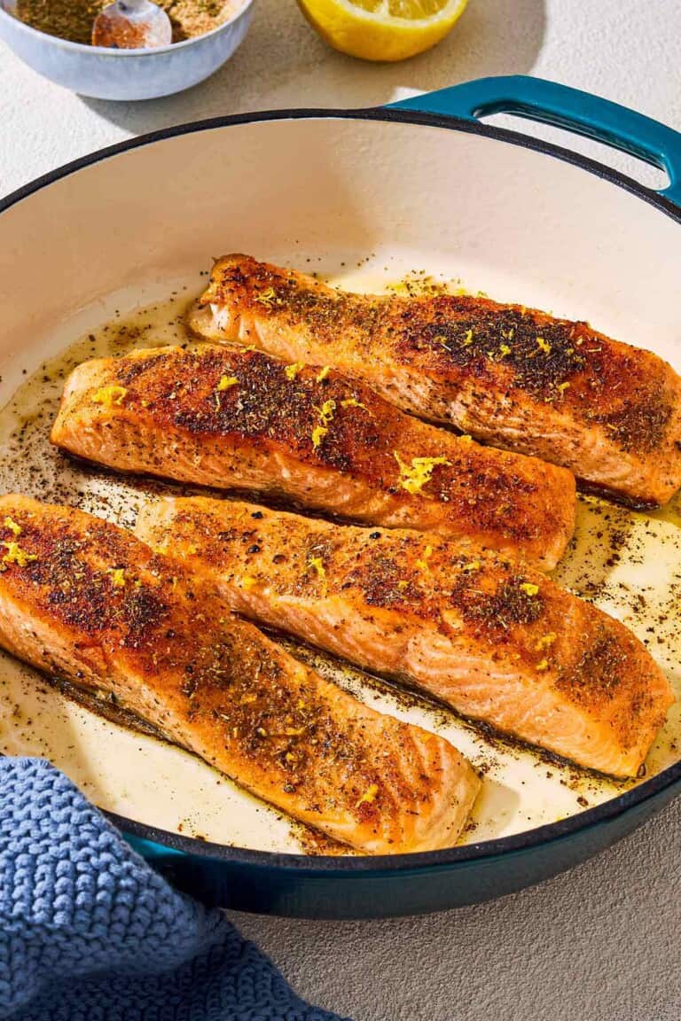 4 salmon filets in a skillet after being pan seared. Next to this is a bowl of spices, a juiced lemon half and a towel.