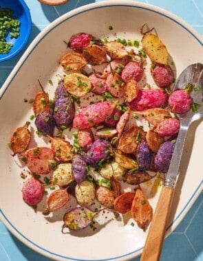 An overhead photo of roasted radishes garnished with chopped chives on a serving platter with a spoon. Next to this is a bowl of chives, and a bowl of the seasonings.