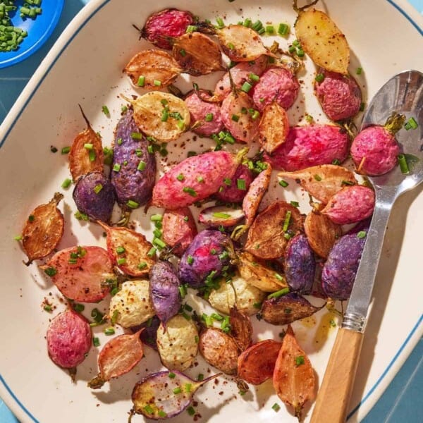 An overhead photo of roasted radishes garnished with chopped chives on a serving platter with a spoon. Next to this is a bowl of chives, and a bowl of the seasonings.