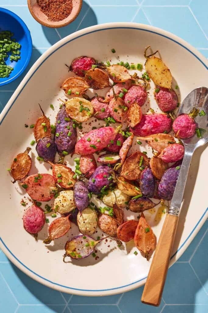 An overhead photo of roasted radishes garnished with chopped chives on a serving platter with a spoon. Next to this is a bowl of chives, and a bowl of the seasonings.