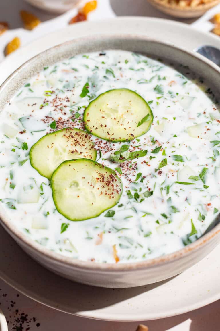 A close up of a bowl of cucumber soup garnished with sumac, parsley and cucumber slices on a plate.
