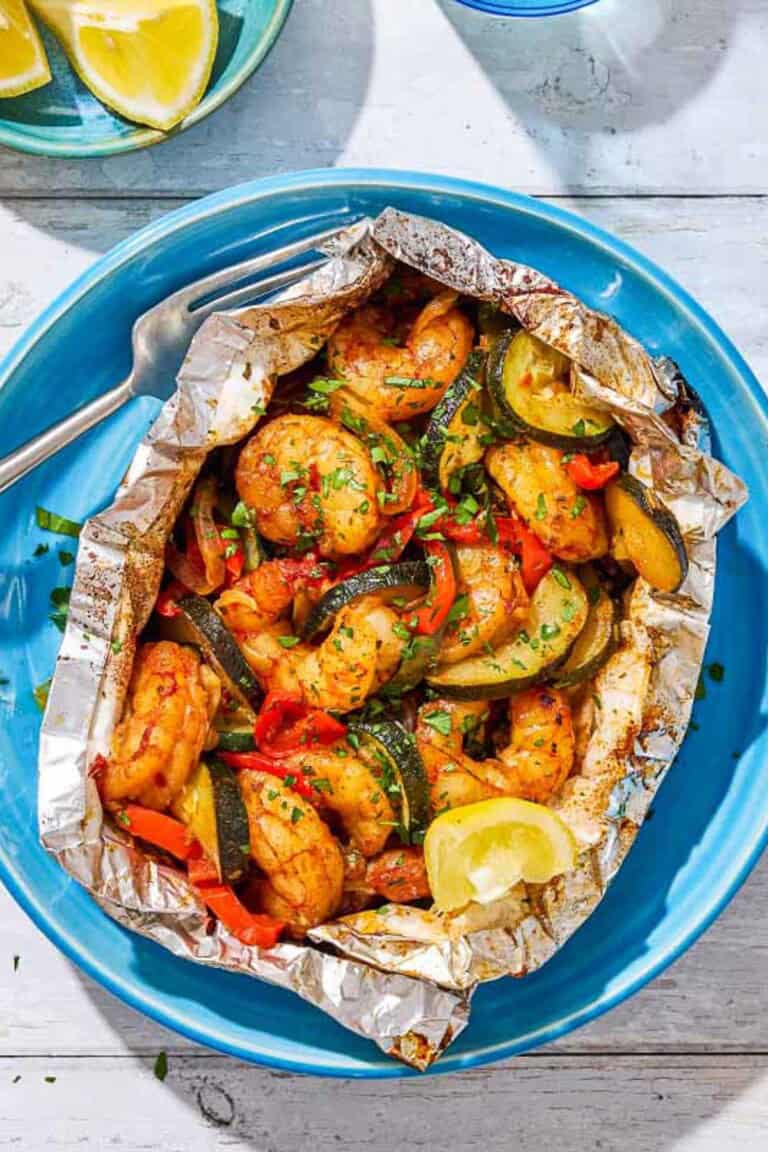 An opened grilled shrimp foil packet with a lemon wedge on a plate with a fork next to a plate of lemon wedges.