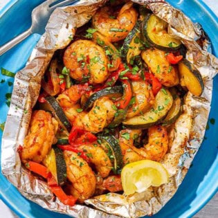 An opened grilled shrimp foil packet with a lemon wedge on a plate with a fork.