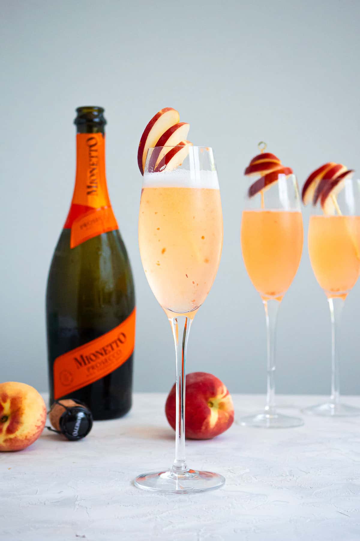 A glass of peach bellini garnished with peach slices in front of a bottle of prosecco, 2 peaches, and 2 more glasses of the bellini.