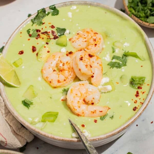 A close up of avocado soup topped with shrimp, cilantro, feta, aleppo pepper and scallions in a bowl with a spoon. Next to this is a lime wedge and bowls of feta and cilantro.