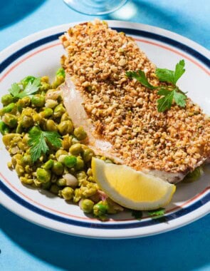 A close up of a serving of baked white fish topped with dukkah on a bed of smashed peas on a plate with a lemon wedge. Next to this is a fork.