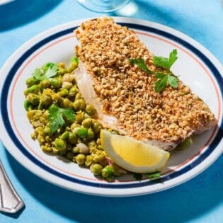 A close up of a serving of baked white fish topped with dukkah on a bed of smashed peas on a plate with a lemon wedge. Next to this is a fork.