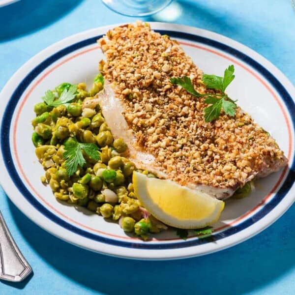 A close up of a serving of baked white fish topped with dukkah on a bed of smashed peas on a plate with a lemon wedge. Next to this is a fork.