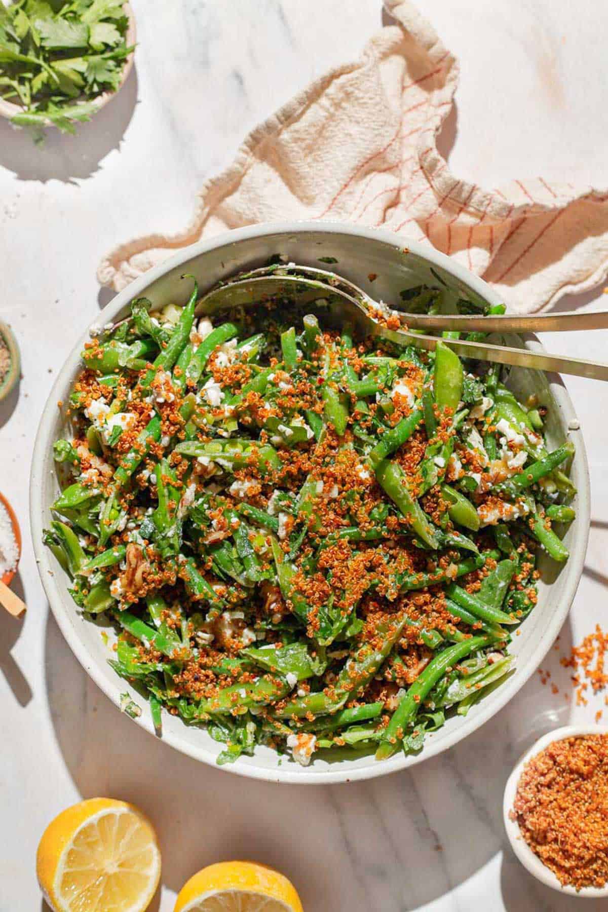An overhead photo of green bean and snap pea salad with crispy quinoa in a bowl with serving utensils. Next to this is a kitchen towel, two lemon halves, and bowls of parsley and crispy quinoa.
